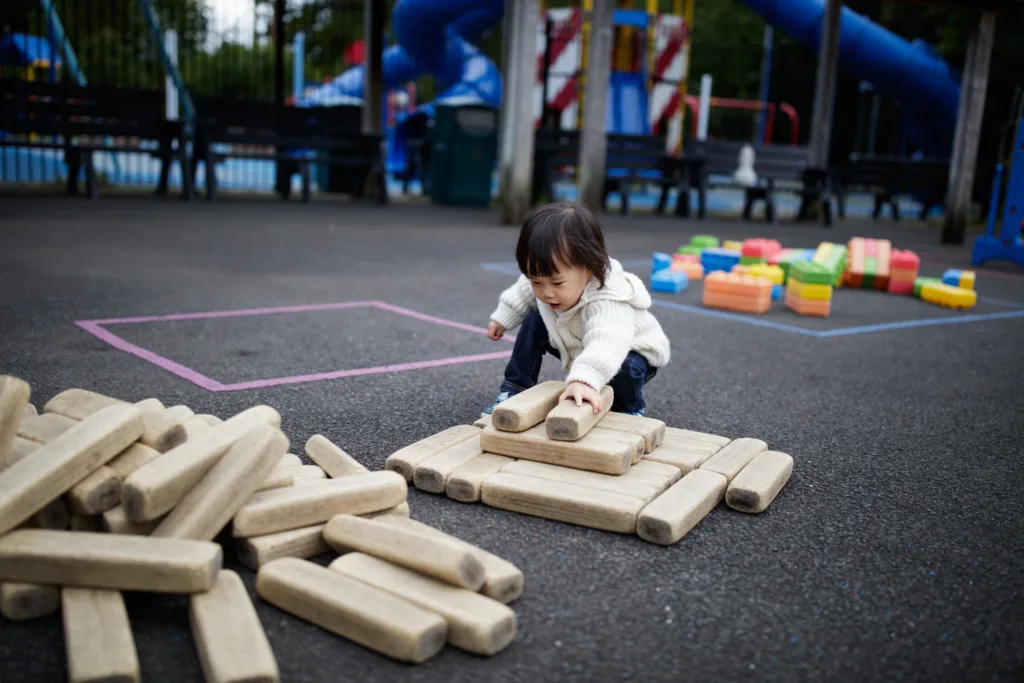 An infant toddler crouches on an outdoor playground, actively building a structure by arranging large, rectangular wooden blocks on the asphalt ground.