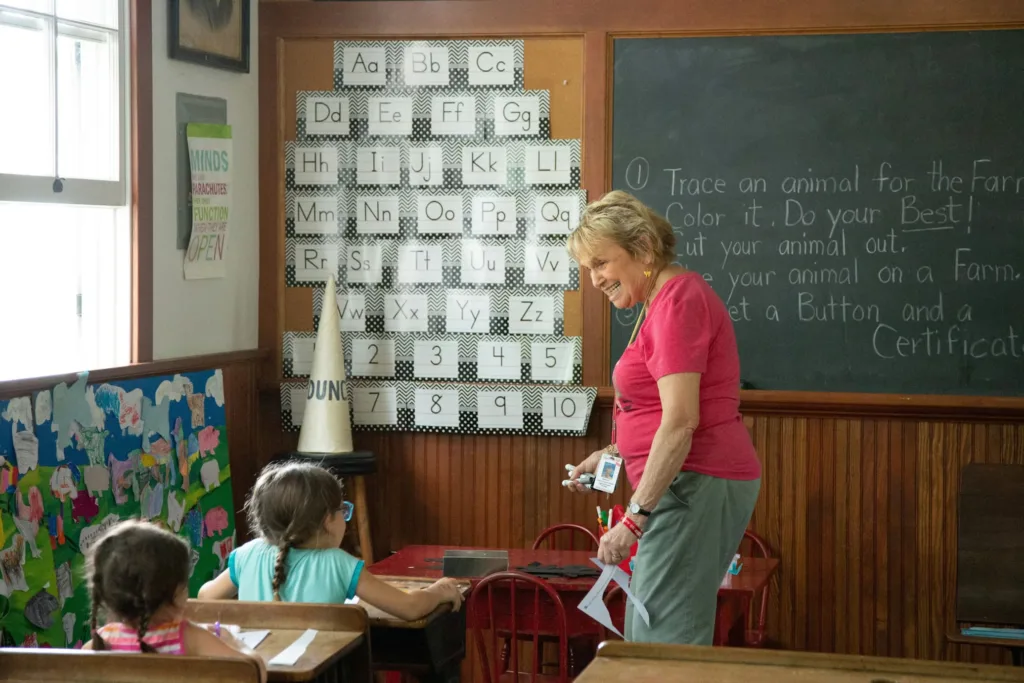 A friendly teacher in preschool stands smiling in front of an alphabet chart, holding cut-out shapes for children seated at small desks in a bright classroom. This scene captures the caring and educational environment that helps young children develop early learning skills and social connections.