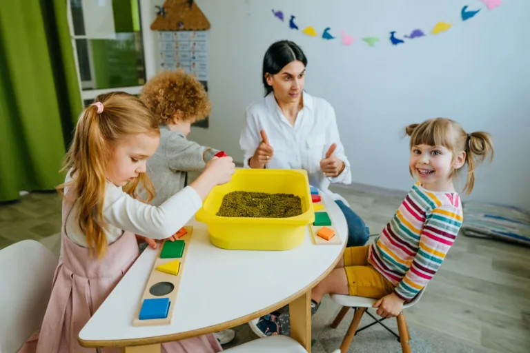 A caregiver at a home daycare gives a thumbs-up to a group of young children actively engaged with shape puzzles and a sensory bin filled with green lentils on a small wooden table.