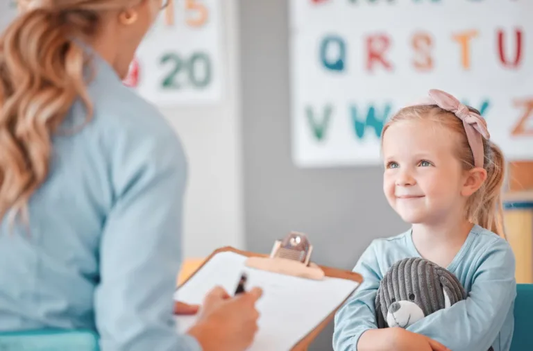 A dedicated teacher sitting at a low wooden table, warmly guiding two toddlers as they sort colorful geometric shapes and practice fine motor tasks inside a bright, licensed central city junior kindergarten classroom.